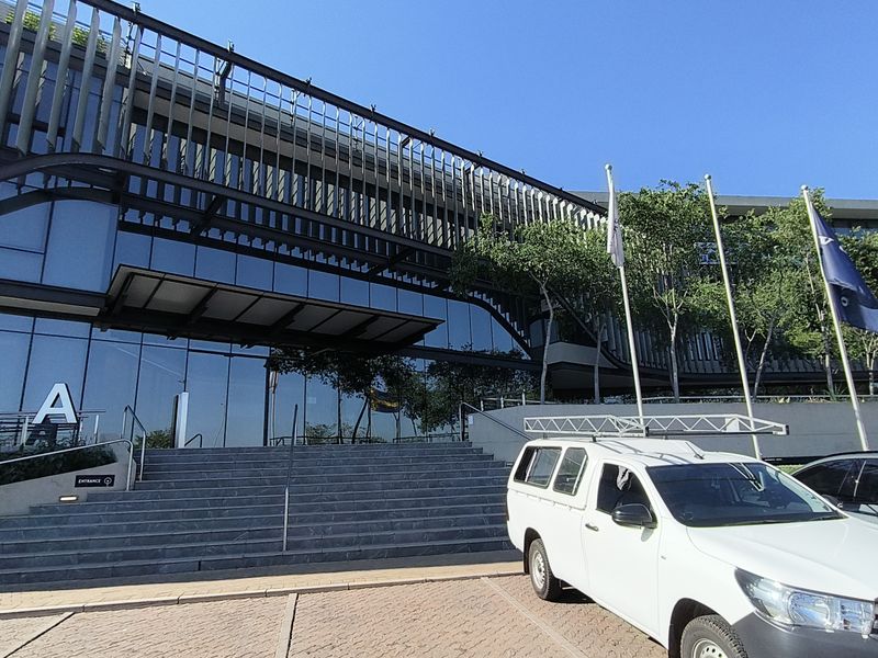 Modern office building entrance featuring a glass curtain wall facade, architectural louvred sun shading, and wide stone steps leading to the main entry.