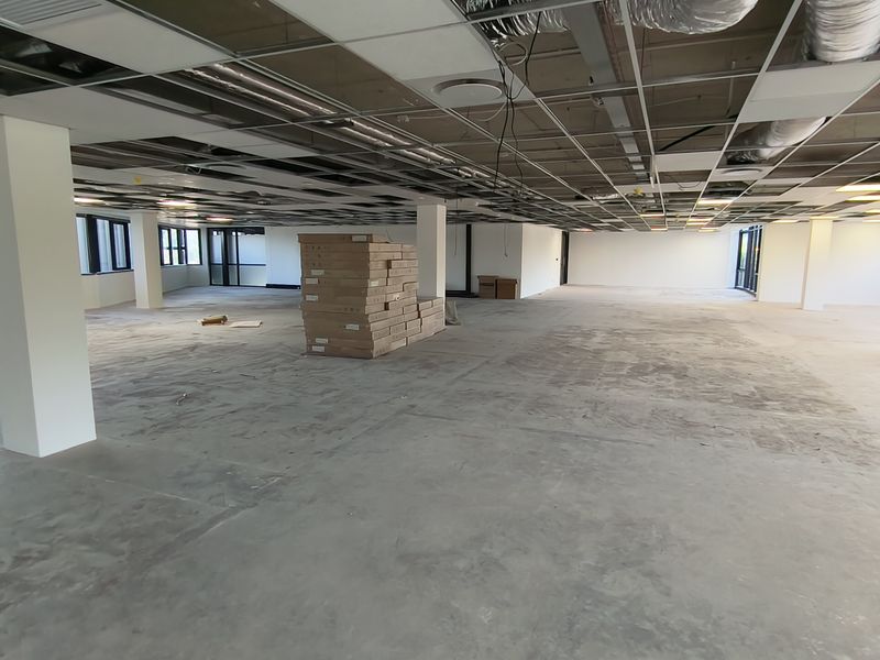 A large, open-plan office space currently under renovation, featuring exposed ceiling infrastructure, structural columns, and expansive windows providing natural light.