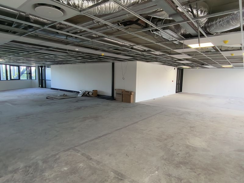 An expansive, open-plan office space currently under renovation, featuring exposed ceiling infrastructure, large windows, and unfinished concrete flooring.