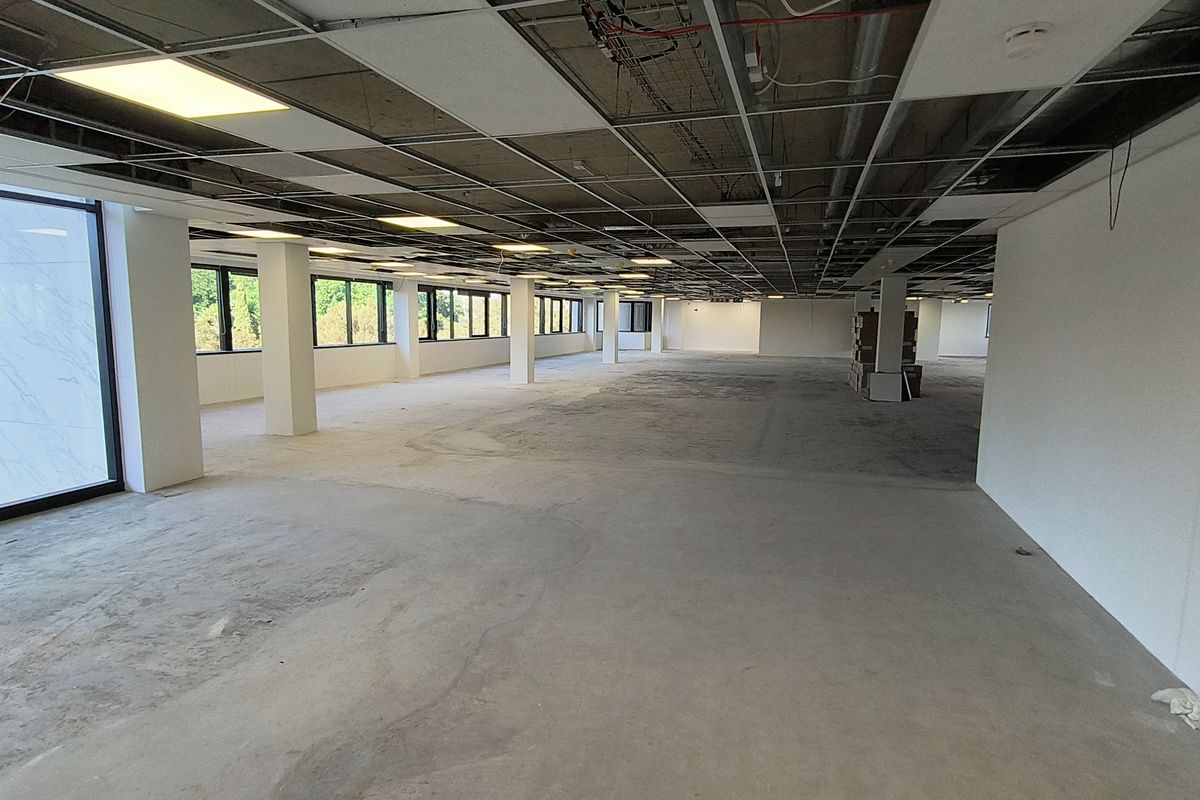 A large, open-plan office space currently in shell condition with exposed ceiling grid, concrete flooring, and extensive natural light from perimeter windows.