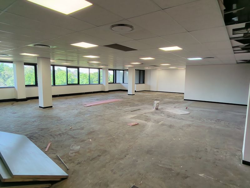 A large, open-plan office space currently undergoing renovation, featuring a suspended ceiling, recessed lighting, and expansive windows providing natural light.