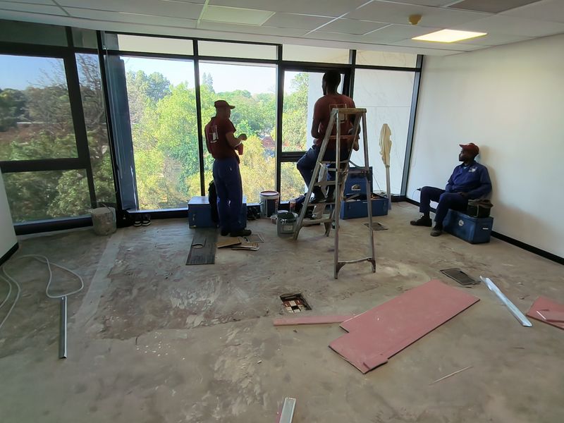 An office space currently undergoing renovations, featuring large floor-to-ceiling windows with natural light and an unfinished floor surface.