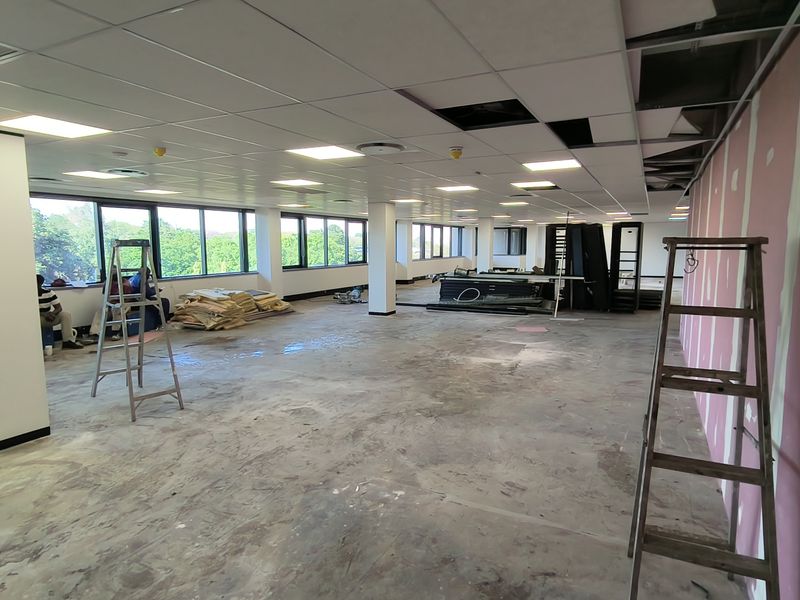 An open-plan office space currently undergoing renovations, featuring large windows, a suspended ceiling grid, and unfinished wall and floor surfaces.