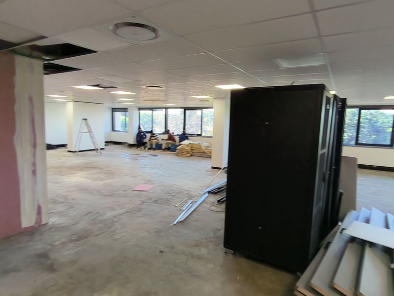 An open-plan office space currently undergoing renovations, featuring large windows, a suspended ceiling grid, and exposed wall surfaces.