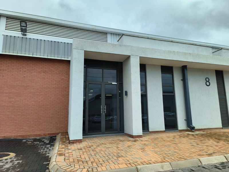Front entrance of an industrial unit featuring a modern glass double door, brick and concrete facade, and paved exterior access.