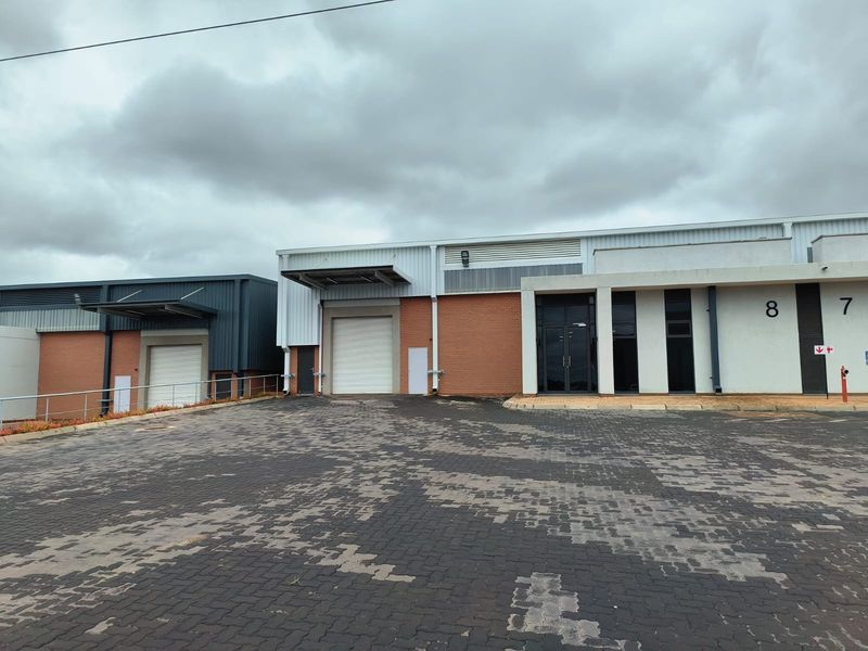Front exterior view of an industrial warehouse complex featuring roller shutter doors, brick and metal cladding, and a paved loading area.