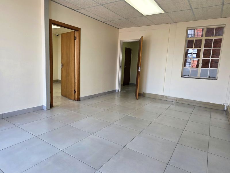 A vacant, well-lit office space featuring neutral tiled flooring, a suspended ceiling with recessed lighting, and multiple access points through wooden doors.