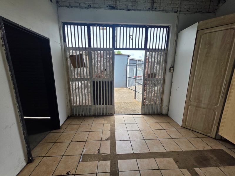 An interior view of a utility space featuring tiled flooring, a metal security gate leading to an outdoor area, and exposed brickwork.