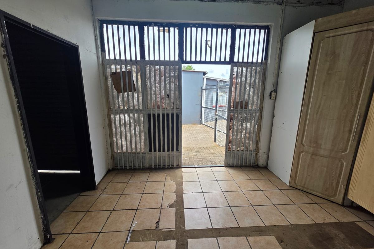 An interior view of a utility space featuring tiled flooring, a metal security gate leading to an outdoor area, and exposed brickwork.