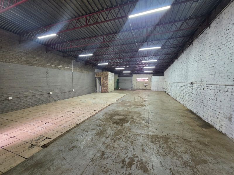 Spacious industrial warehouse or workshop area featuring exposed steel roof trusses, concrete flooring, painted brick walls, and overhead fluorescent lighting.