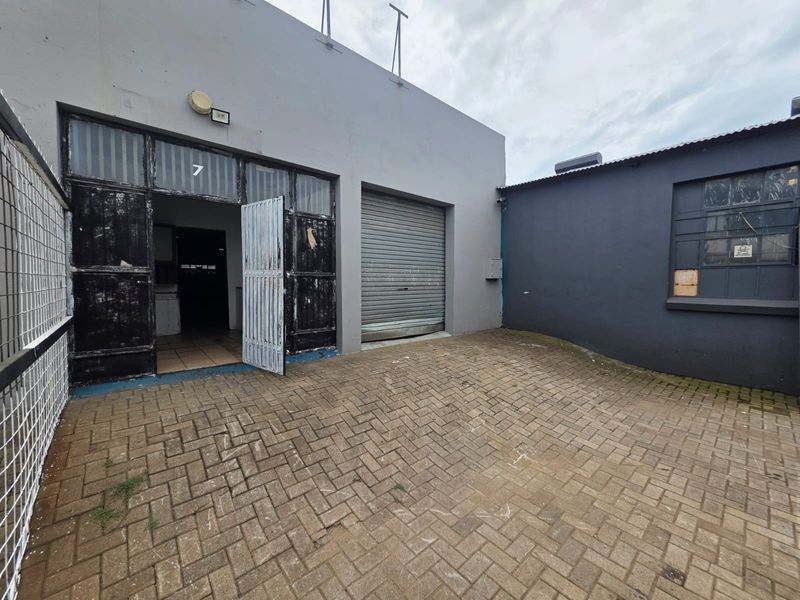 Exterior view of a commercial unit featuring a paved courtyard, roller shutter door, and metal-framed entrance.