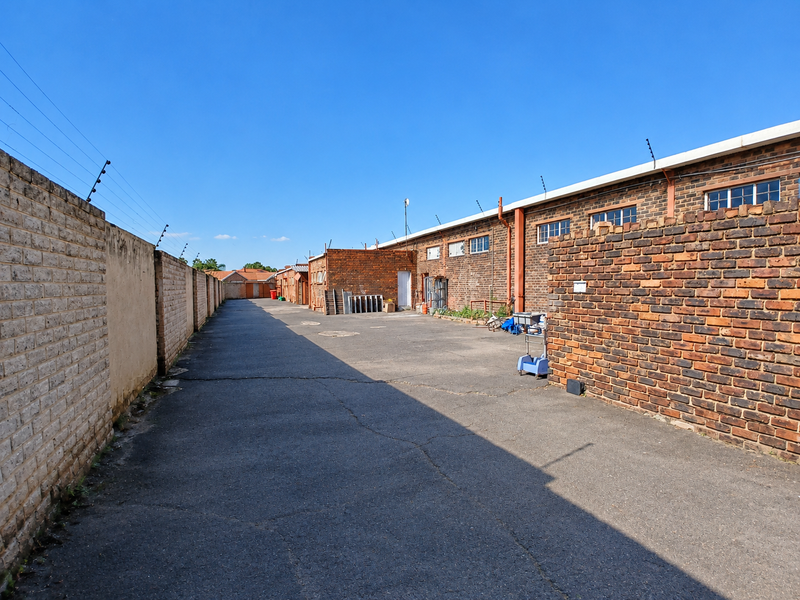 A rear exterior view of a commercial property featuring an asphalt driveway, brick boundary walls with electric fencing, and exposed brick building facades.