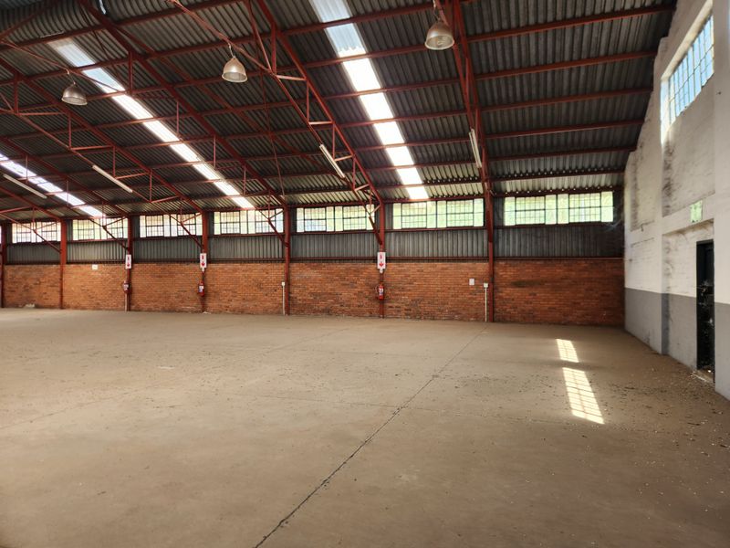 Spacious industrial warehouse featuring high ceilings with exposed steel trusses, abundant natural light from skylights, concrete flooring, and exposed brick walls.