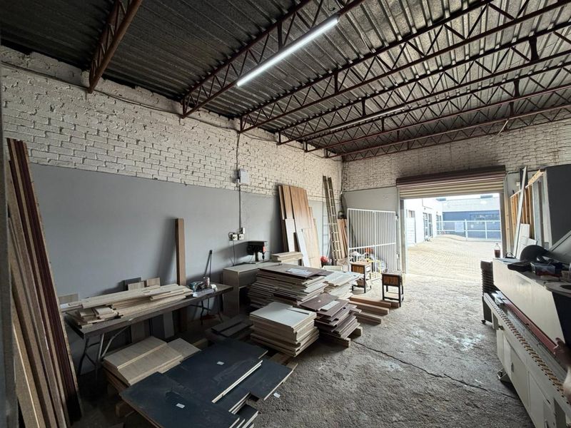 Industrial workshop space featuring high exposed steel truss ceilings, concrete flooring, and a large roller shutter door providing direct access to the exterior yard.