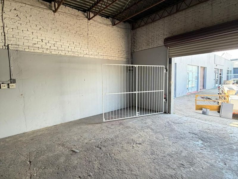 Industrial warehouse space featuring high exposed steel truss ceilings, painted brick walls, concrete flooring, and a large roller shutter door for vehicle access.