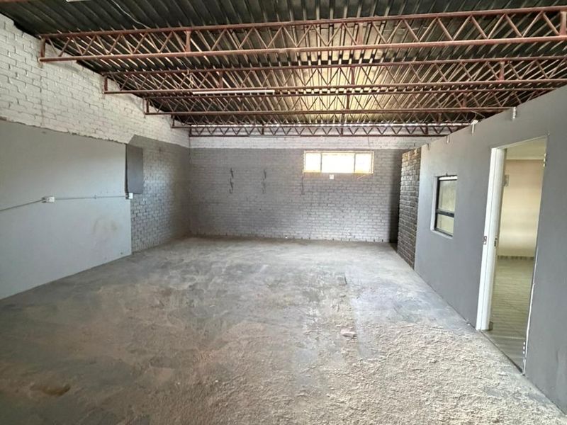 An open-plan industrial space featuring exposed steel roof trusses, painted brick walls, and a raw concrete floor, suitable for workshop or storage use.