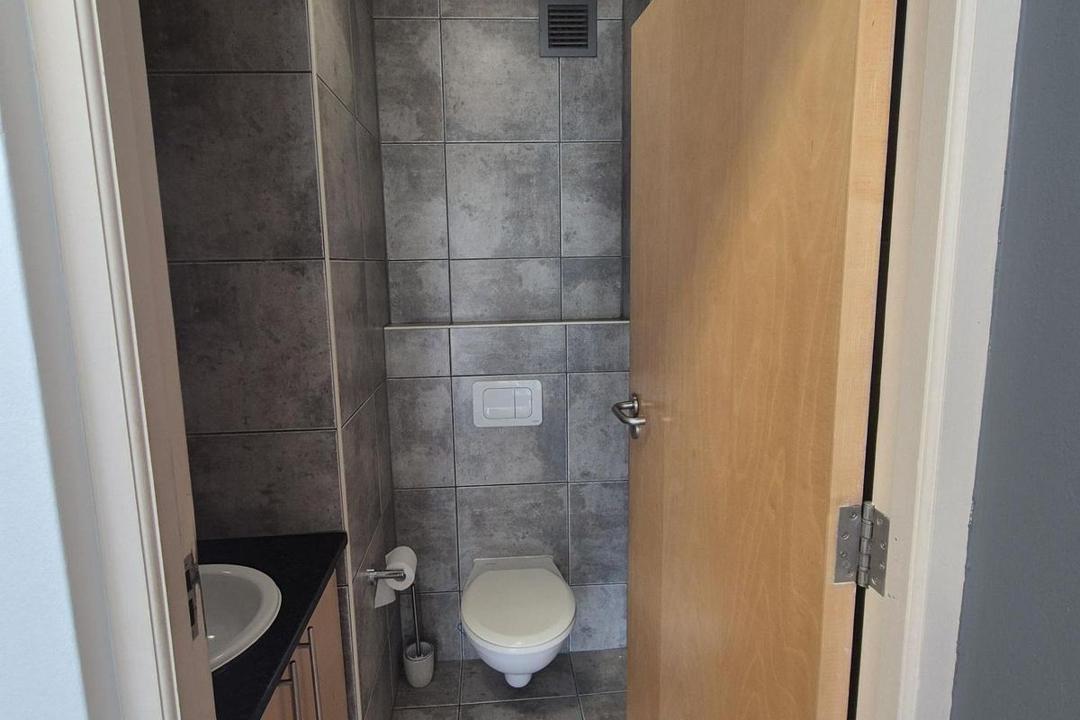 A compact, modern office restroom featuring grey tiled walls and flooring, a wall-mounted toilet, and a built-in vanity cabinet with a sink.