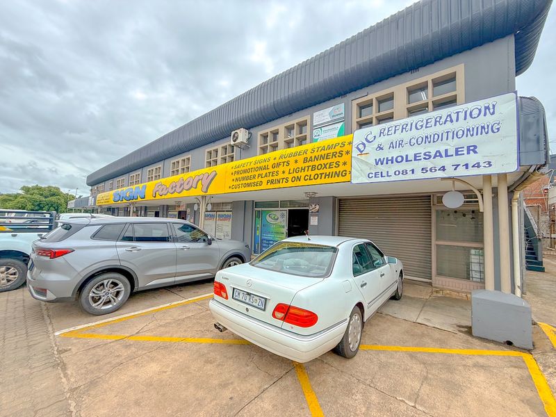 Exterior view of an industrial commercial property featuring multiple business units with roller shutter doors and paved parking area.