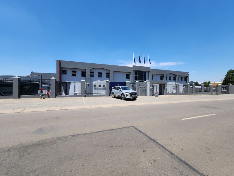 Front elevation of a modern industrial facility featuring a secure perimeter fence, paved driveway, and professional facade with flagpoles.
