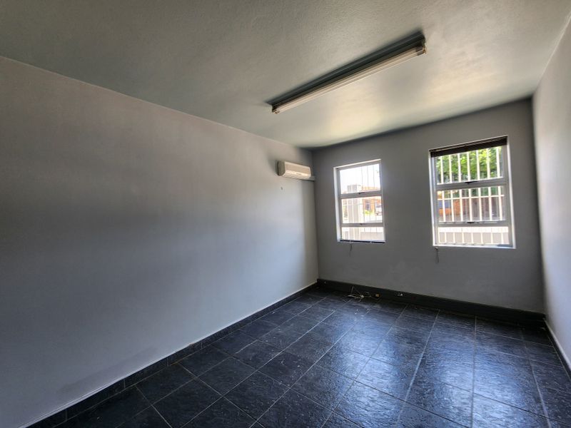 A clean, empty office space featuring tiled flooring, security bars on the windows, and a wall-mounted air conditioning unit.