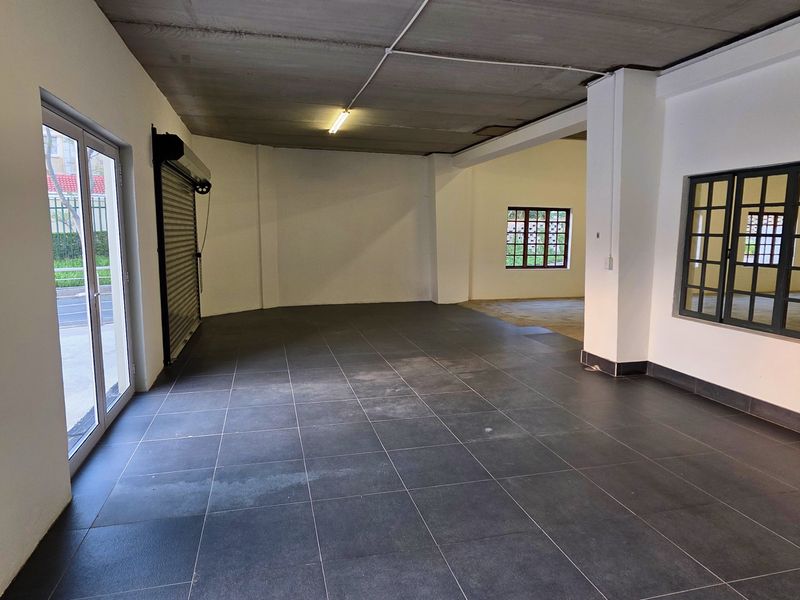 Spacious industrial utility area featuring dark tiled flooring, exposed concrete ceilings, and large grid-style windows with a secure roller shutter door entrance.