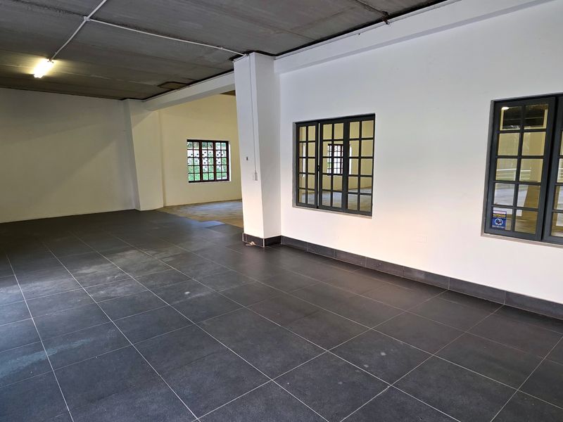 Spacious industrial utility area featuring dark tiled flooring, exposed concrete ceiling, and large grid-pattern windows, offering a clean and versatile open-plan workspace.