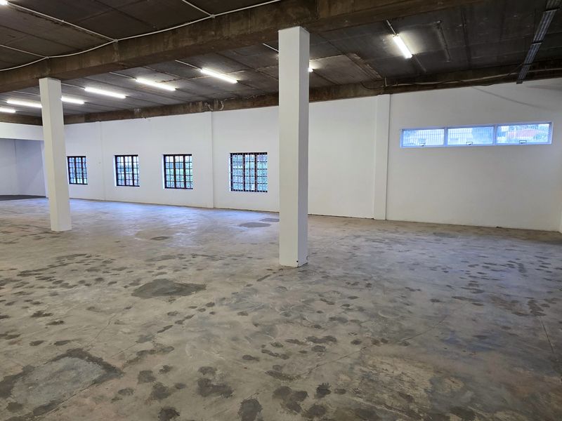 Spacious open-plan industrial warehouse area featuring exposed concrete ceilings, structural columns, security-barred windows, and a roller shutter door.