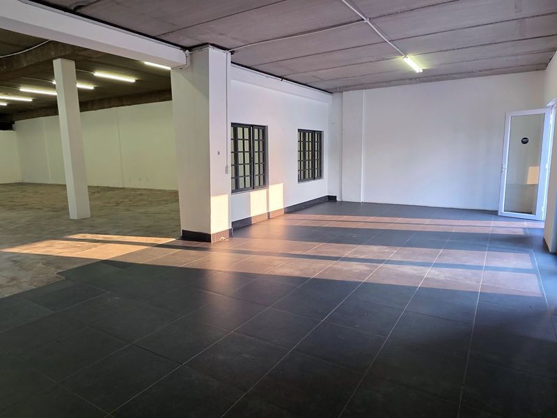 Spacious open-plan industrial unit featuring a mix of tiled and concrete flooring, structural columns, fluorescent lighting, and a secure roller shutter door.
