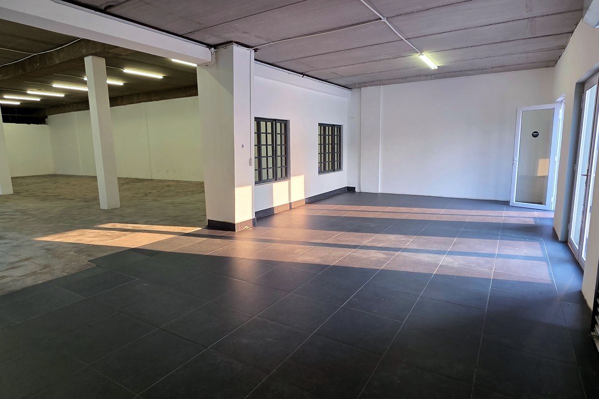 Spacious open-plan industrial unit featuring a mix of tiled and concrete flooring, structural columns, fluorescent lighting, and a secure roller shutter door.