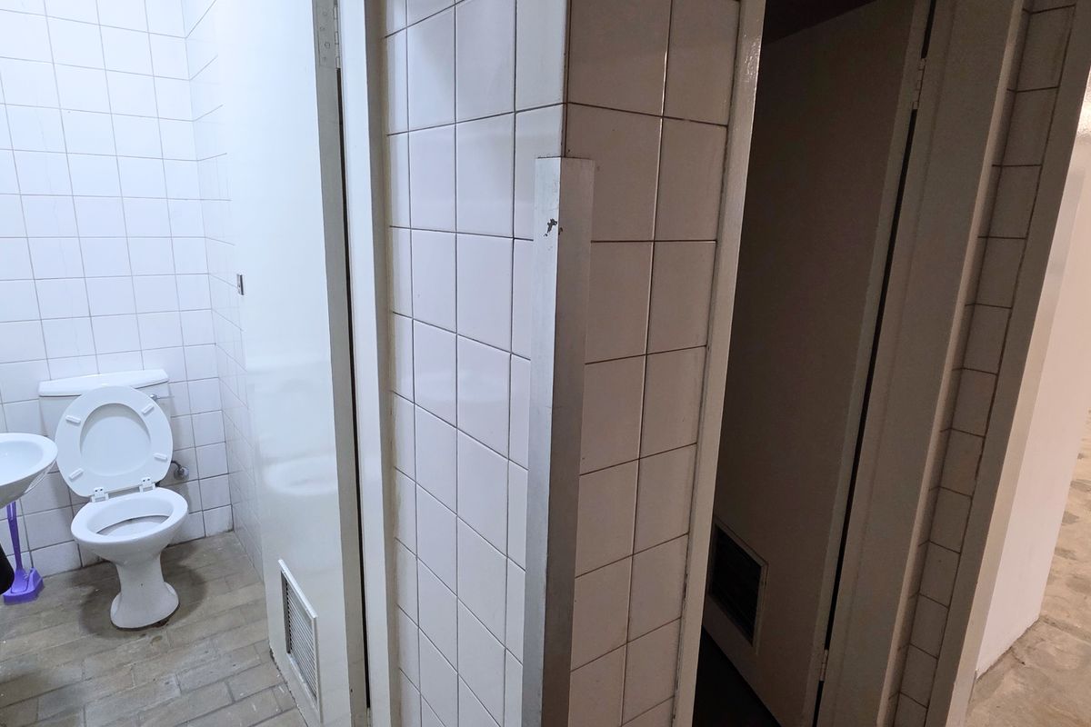 A functional industrial-style bathroom featuring white wall tiles, a wall-mounted basin, and a toilet, adjacent to an open-plan warehouse or office space.