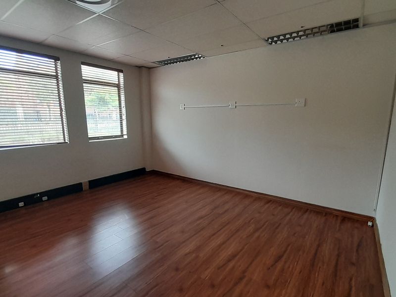 A vacant office space featuring laminate wood flooring, a suspended ceiling with recessed lighting, and large windows with fitted venetian blinds.
