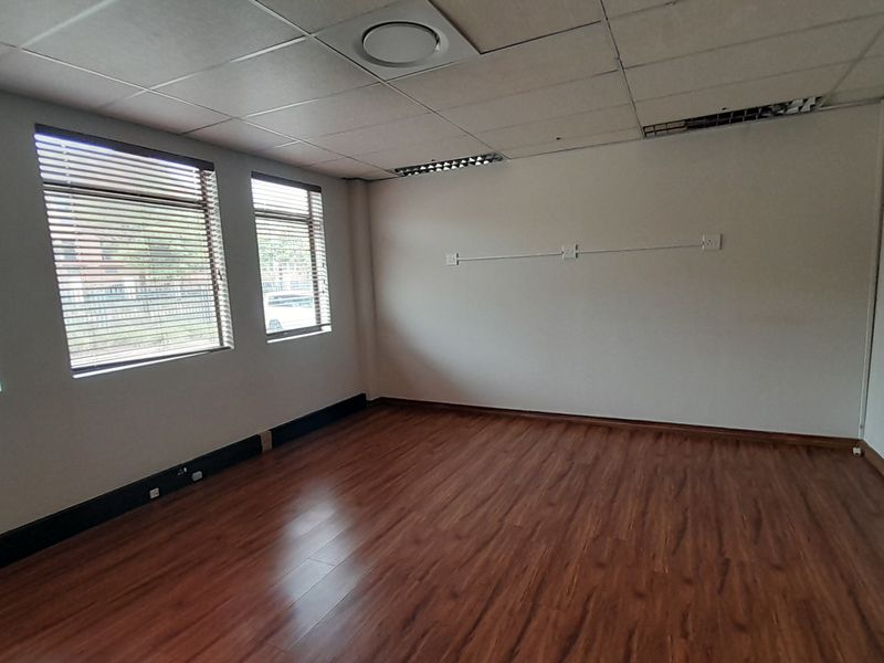 A vacant office space featuring laminate flooring, suspended ceilings with recessed lighting, and large windows fitted with venetian blinds.