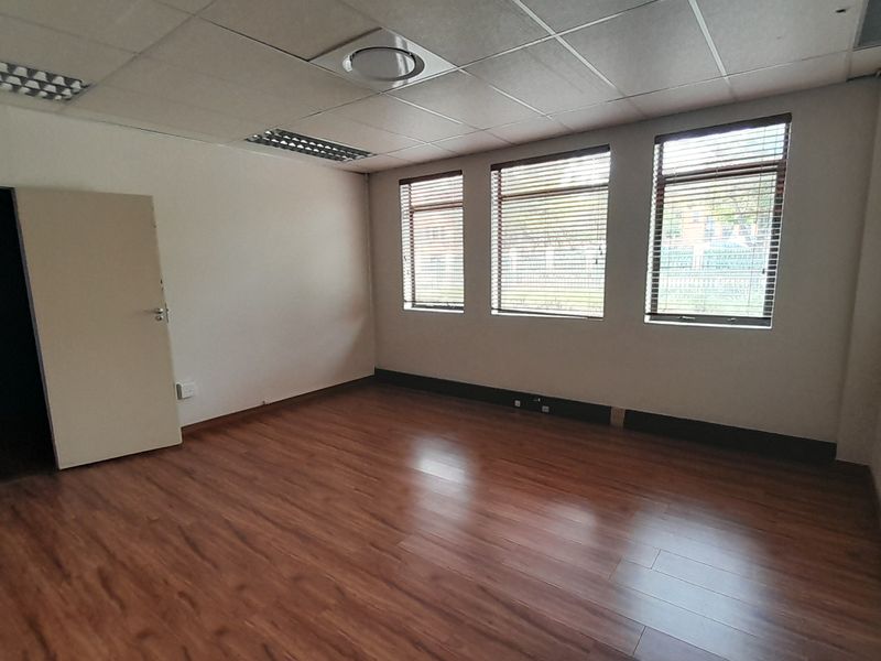 A vacant office space featuring laminate flooring, a suspended ceiling with recessed lighting, and large windows fitted with venetian blinds.