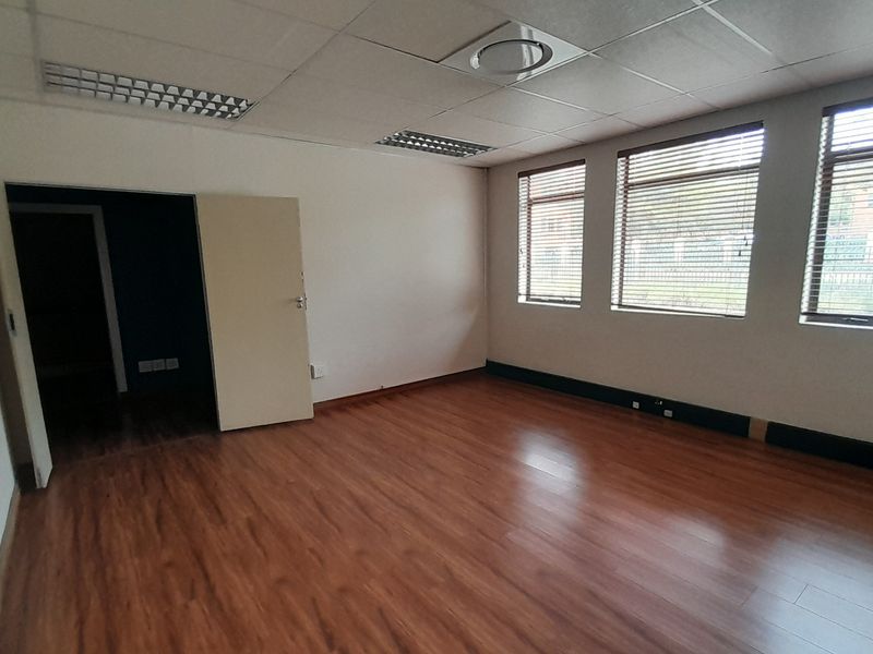 A vacant office space featuring laminate flooring, a suspended ceiling with recessed lighting, and large windows fitted with venetian blinds.