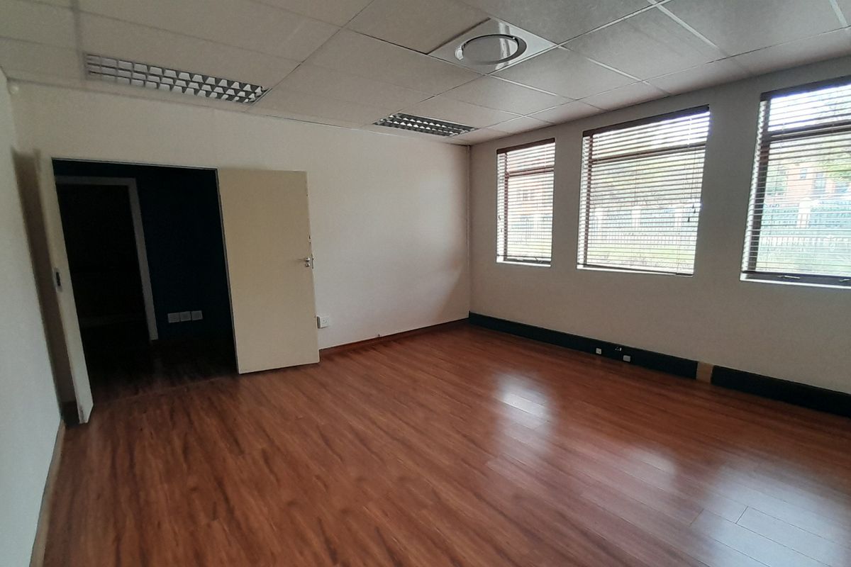 A vacant office space featuring laminate flooring, a suspended ceiling with recessed lighting, and large windows fitted with venetian blinds.