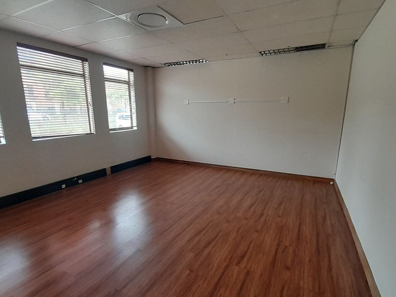 A bright, empty office space featuring laminate wood flooring, a suspended ceiling with recessed lighting, and large windows with fitted venetian blinds.