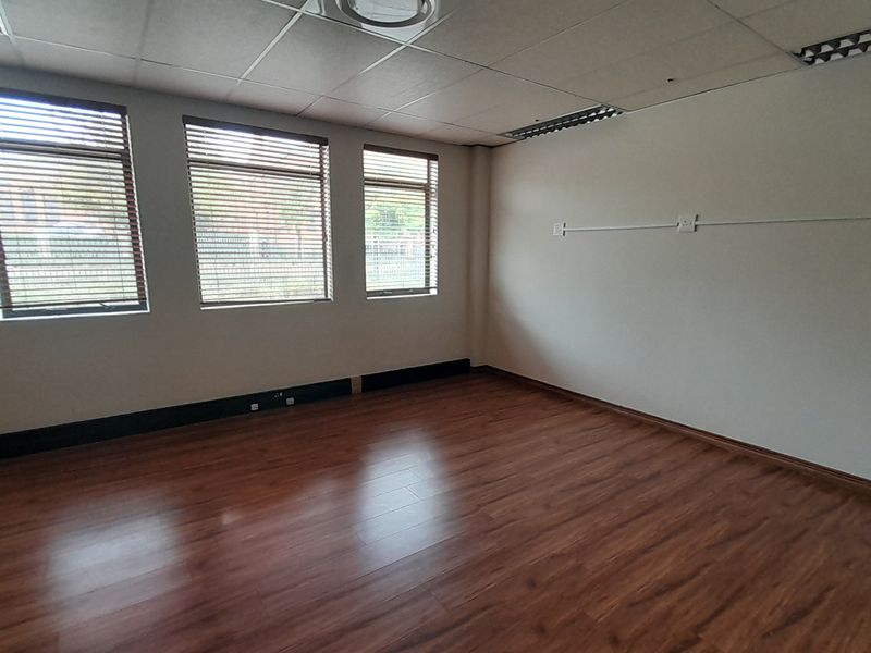 A bright, empty office space featuring laminate flooring, a suspended ceiling with recessed lighting, and large windows fitted with venetian blinds.
