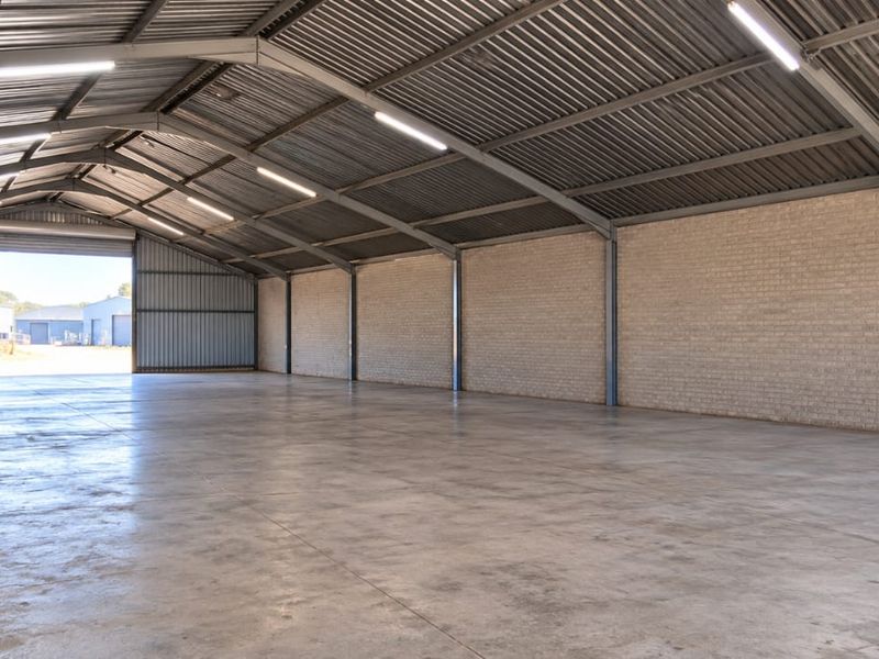 Spacious industrial warehouse featuring a concrete floor, exposed brick walls, steel portal frame construction, and high-clearance corrugated metal roofing with integrated lighting.