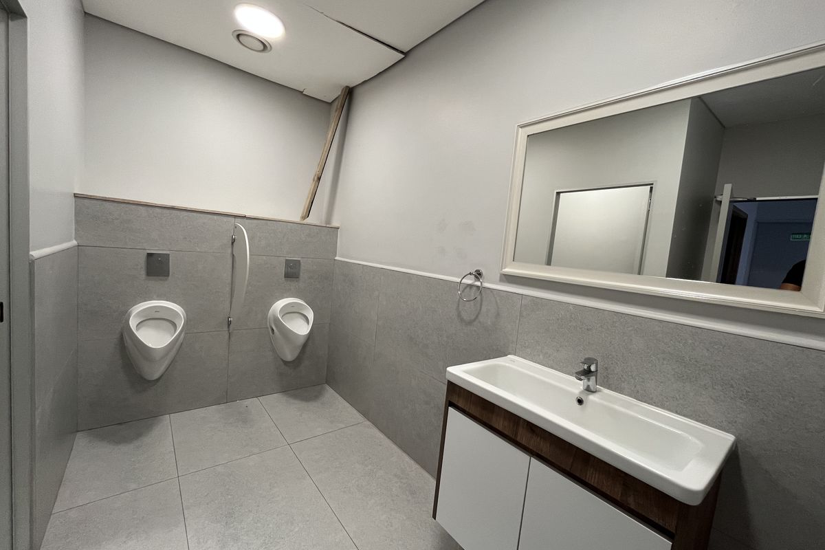 A commercial restroom featuring two wall-mounted urinals, a vanity basin with storage cabinet, and a large wall mirror.
