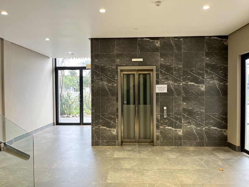 Modern building lobby featuring a stainless steel elevator, dark marble-effect wall cladding, recessed ceiling lighting, and large glass entrance doors.