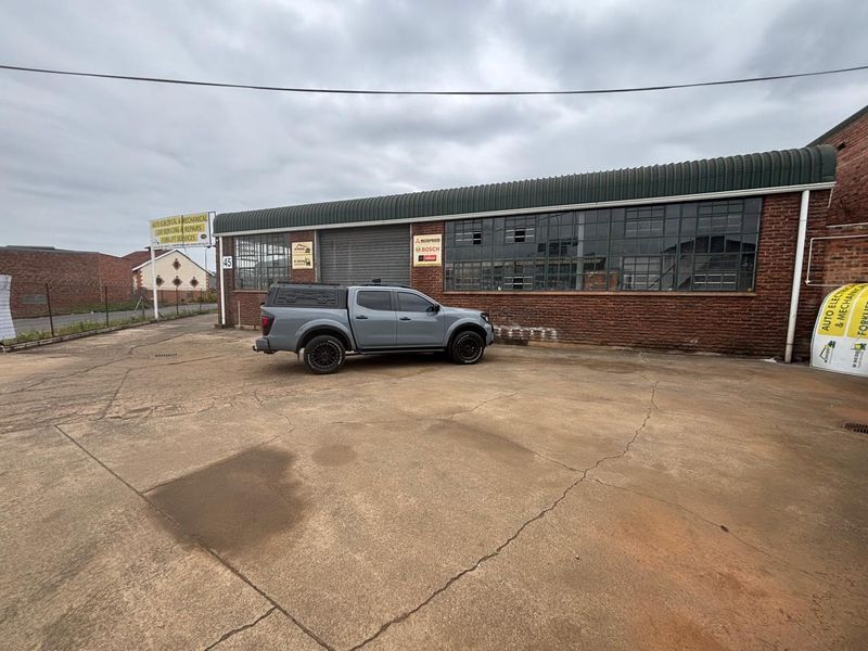 Industrial property exterior featuring a brick facade, large steel-framed windows, a roller shutter door, and a spacious concrete yard area.
