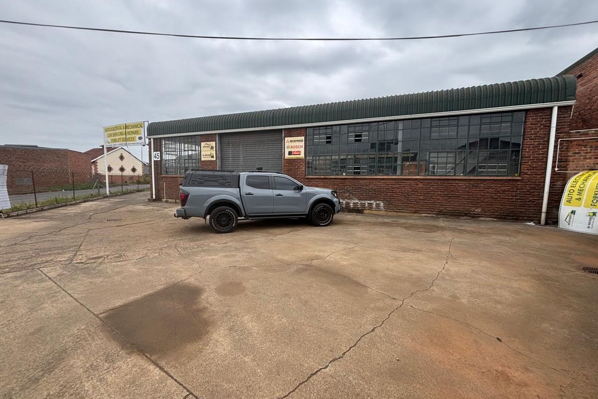 Industrial property exterior featuring a brick facade, large steel-framed windows, a roller shutter door, and a spacious concrete yard area.
