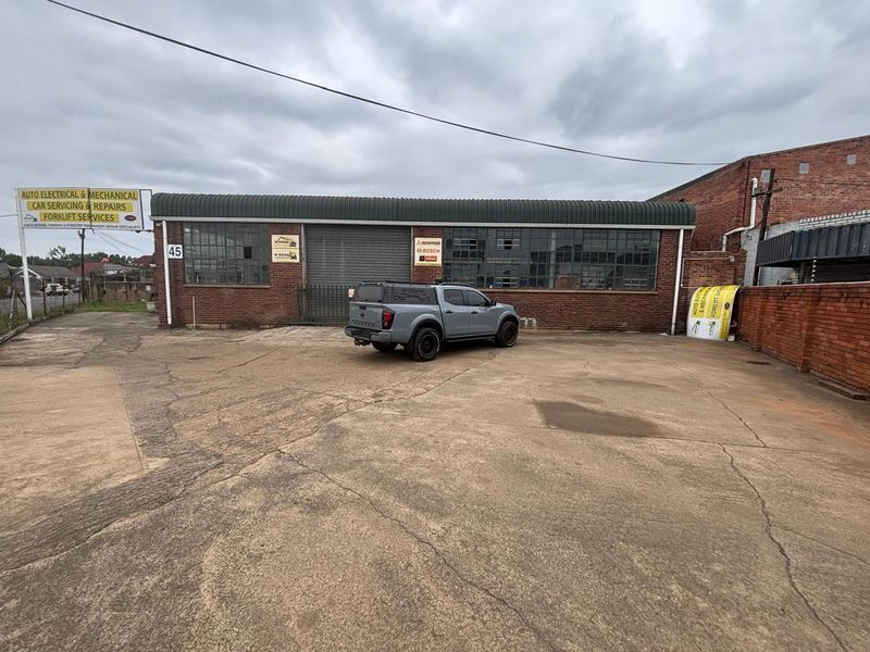 Industrial property exterior featuring a brick facade, large roller shutter door, steel-framed windows, and a spacious concrete driveway area.