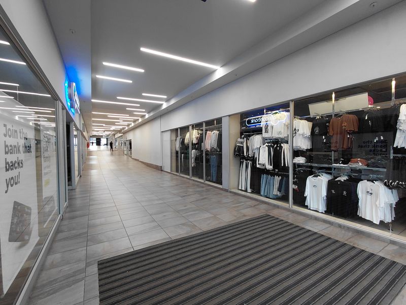 A well-lit commercial hallway featuring tiled flooring, recessed linear ceiling lighting, and glass-fronted retail storefronts.