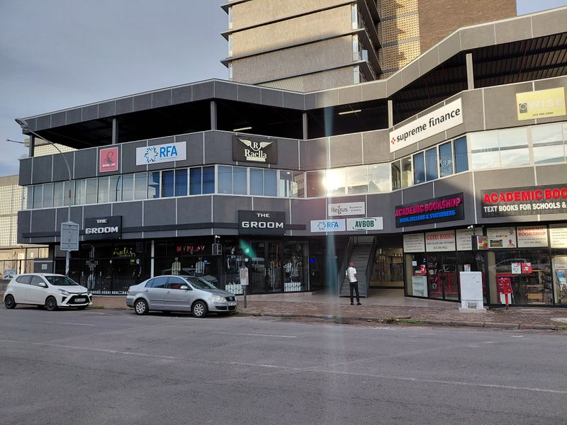 Street-level view of a multi-story commercial building featuring various retail storefronts, office signage, and a central pedestrian access staircase.