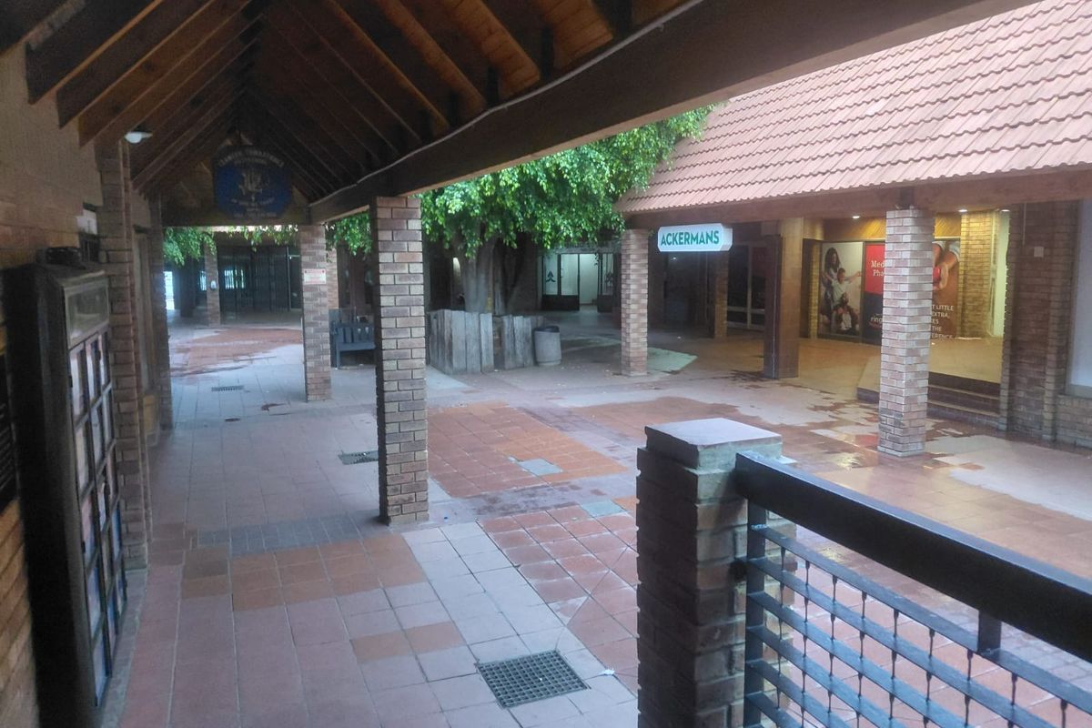 A covered outdoor walkway and courtyard area within a commercial shopping center, featuring brick pillars, tiled flooring, and exposed timber roof trusses.