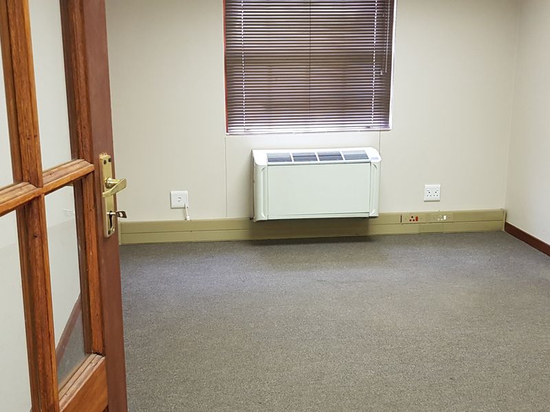 A vacant office space featuring carpeted flooring, a wall-mounted air conditioning unit, and a timber-framed glass entrance door.