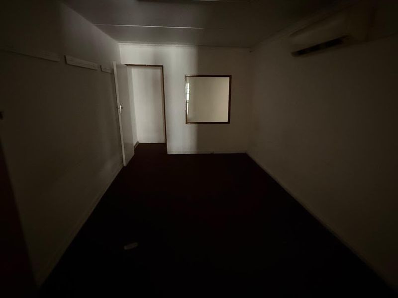 An empty, dimly lit office space featuring a wall-mounted air conditioning unit, a fluorescent light fixture, and an internal window.