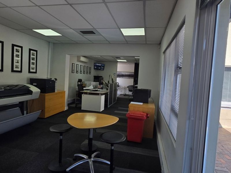 A functional office space featuring a suspended ceiling, recessed lighting, and carpeted flooring, with large windows providing natural light and access to an outdoor area.