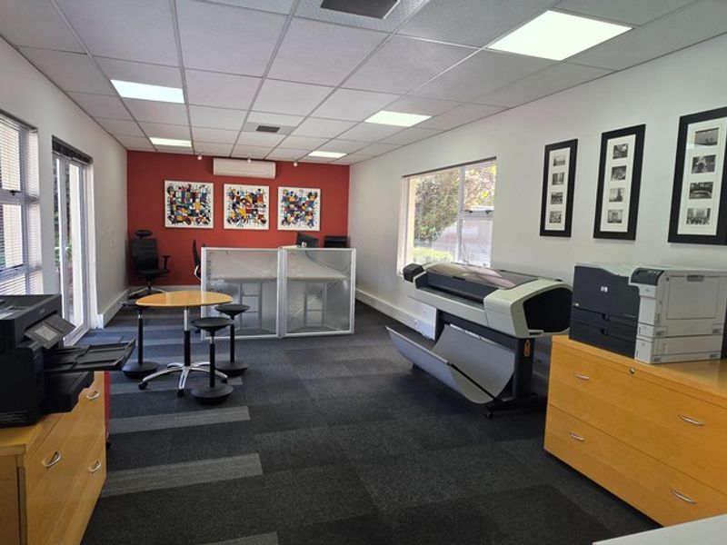 A functional office space featuring carpeted flooring, a suspended ceiling with recessed lighting, and a wall-mounted air conditioning unit.